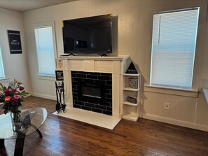 A living room with a fireplace, a flat-screen TV mounted above it, and a glass coffee table. - The Cedar Cottage - Cozy 3BR/1BR Home Near Texas Tech (Lubbock)