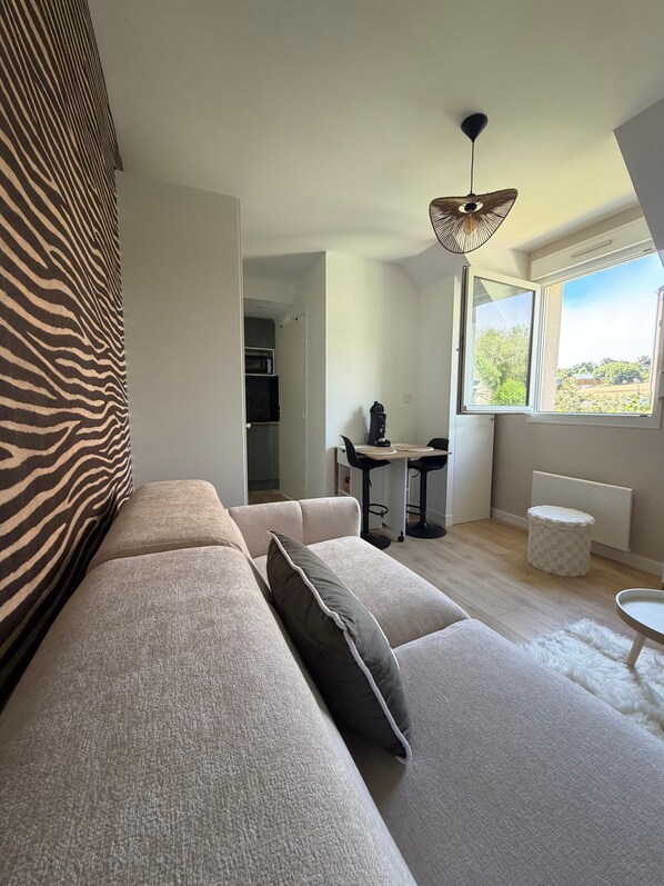 A modern living room with a sofa, a dining table with chairs, a ceiling fan, and a window with a view.