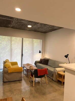 Un salon moderne comprenant un canapé, un fauteuil et une table basse, une table d’appoint en bois et une grande fenêtre avec des stores.
