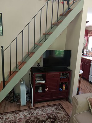A staircase with a wooden finish and metal railings, leading to a room with a television on a wooden stand, a red cabinet with drawers, and a patterned rug on the floor.