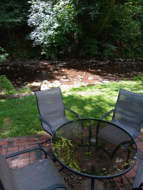 A round glass table with two chairs, a brick pathway, and a stream in the background.
