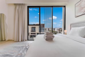 Een hotelkamer met een groot bed, uitzicht op gebouwen door een raam en een stapel handdoeken op het bed.