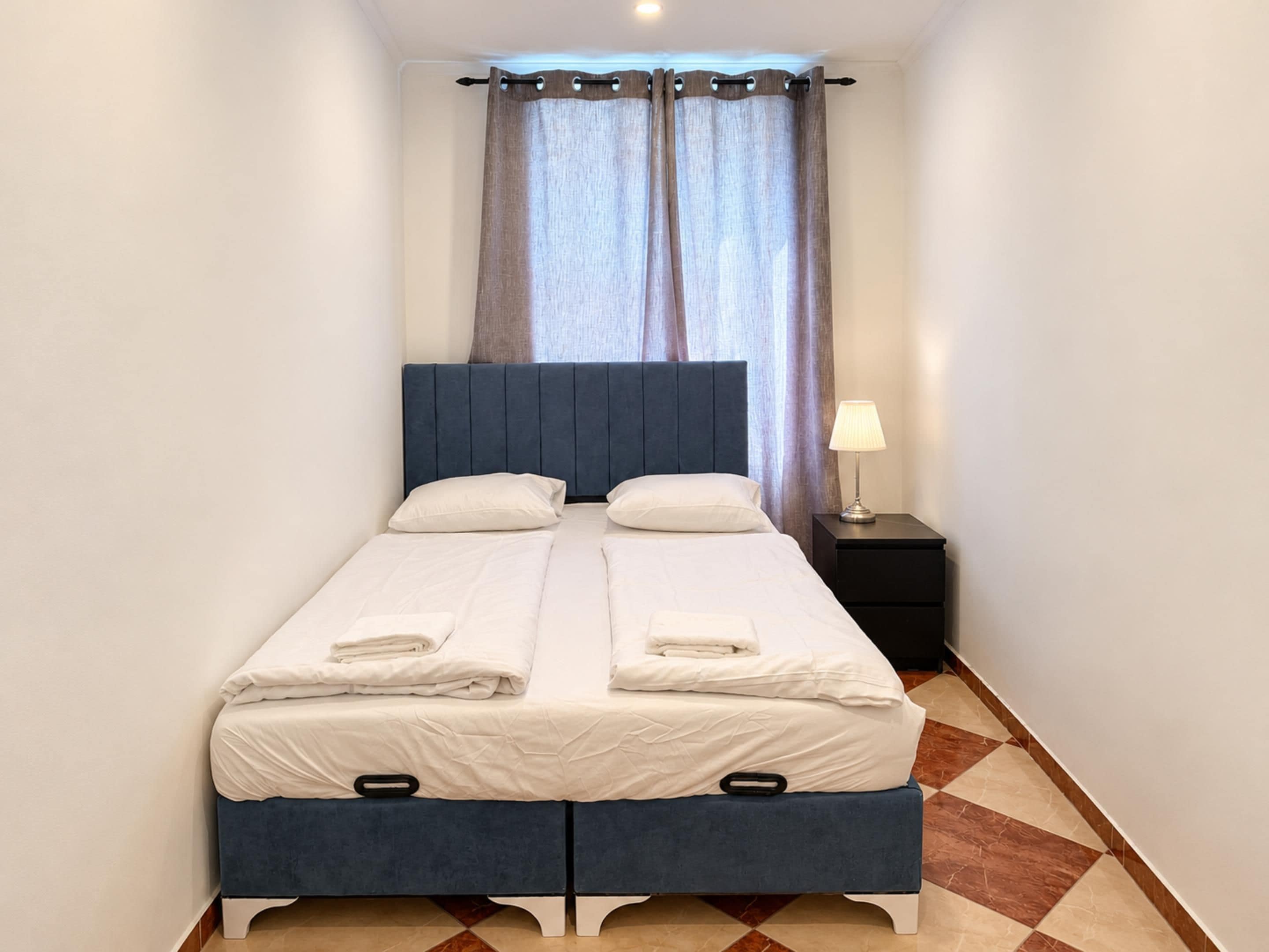 Una cama doble con cabecera, una mesita de noche con lámpara y una ventana con cortinas.
