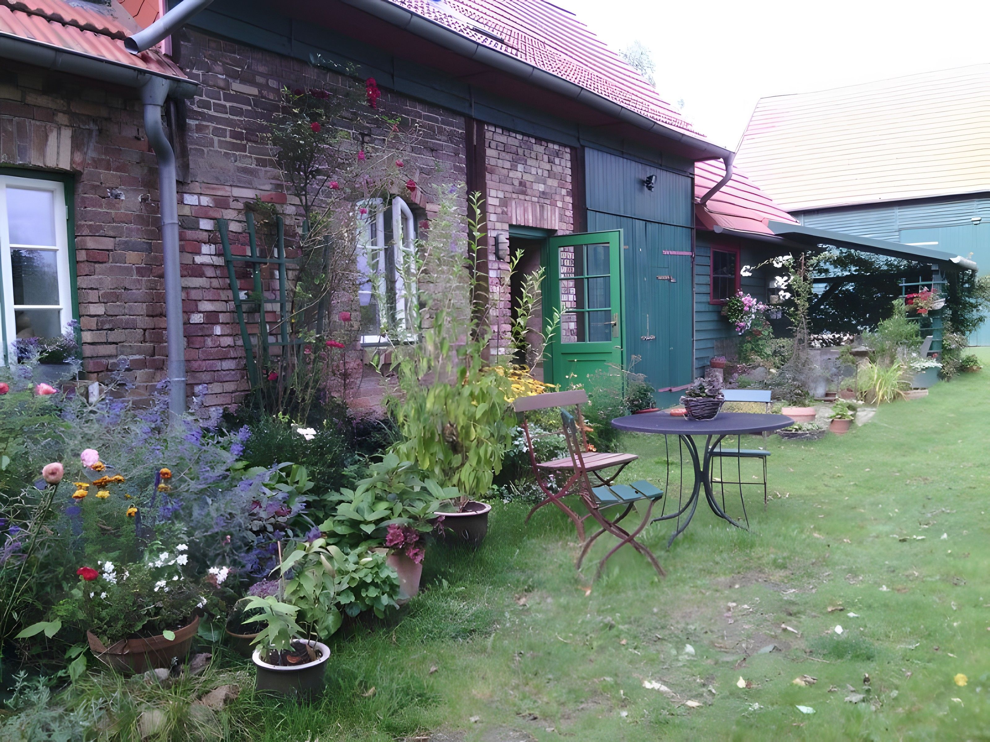 A brick house with a green door, a small garden with various plants and flowers, a table with chairs, and a window with a view of the sky.