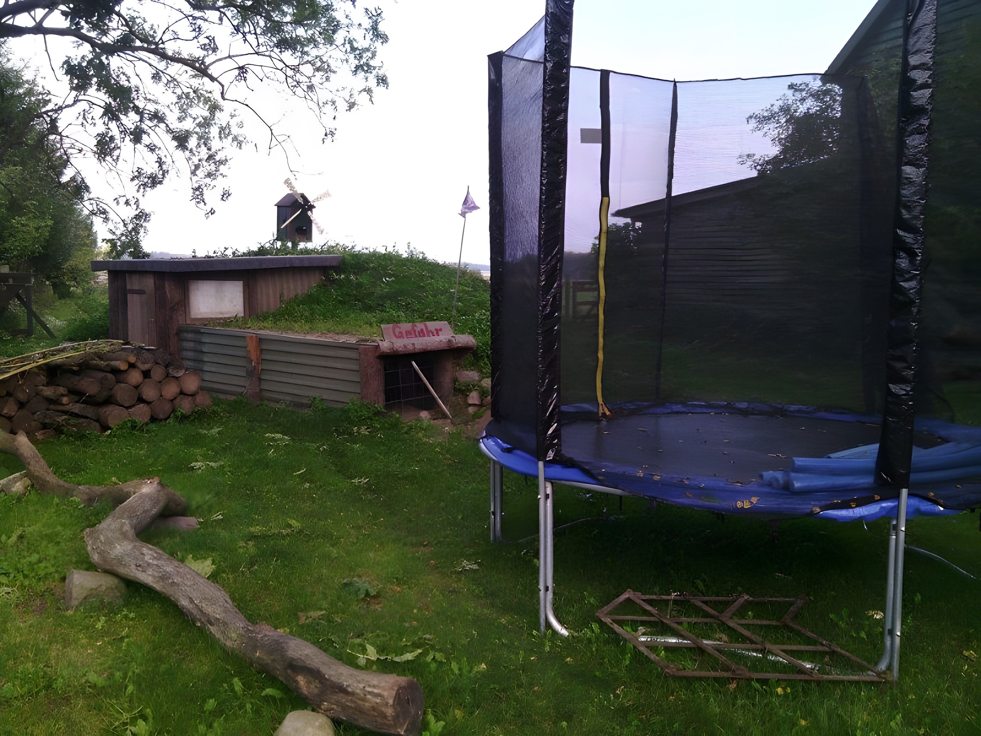 Ein Trampolin mit Sicherheitsnetz, umgeben von einer Rasenfläche mit einer Holzkonstruktion und einem Holzstoß.