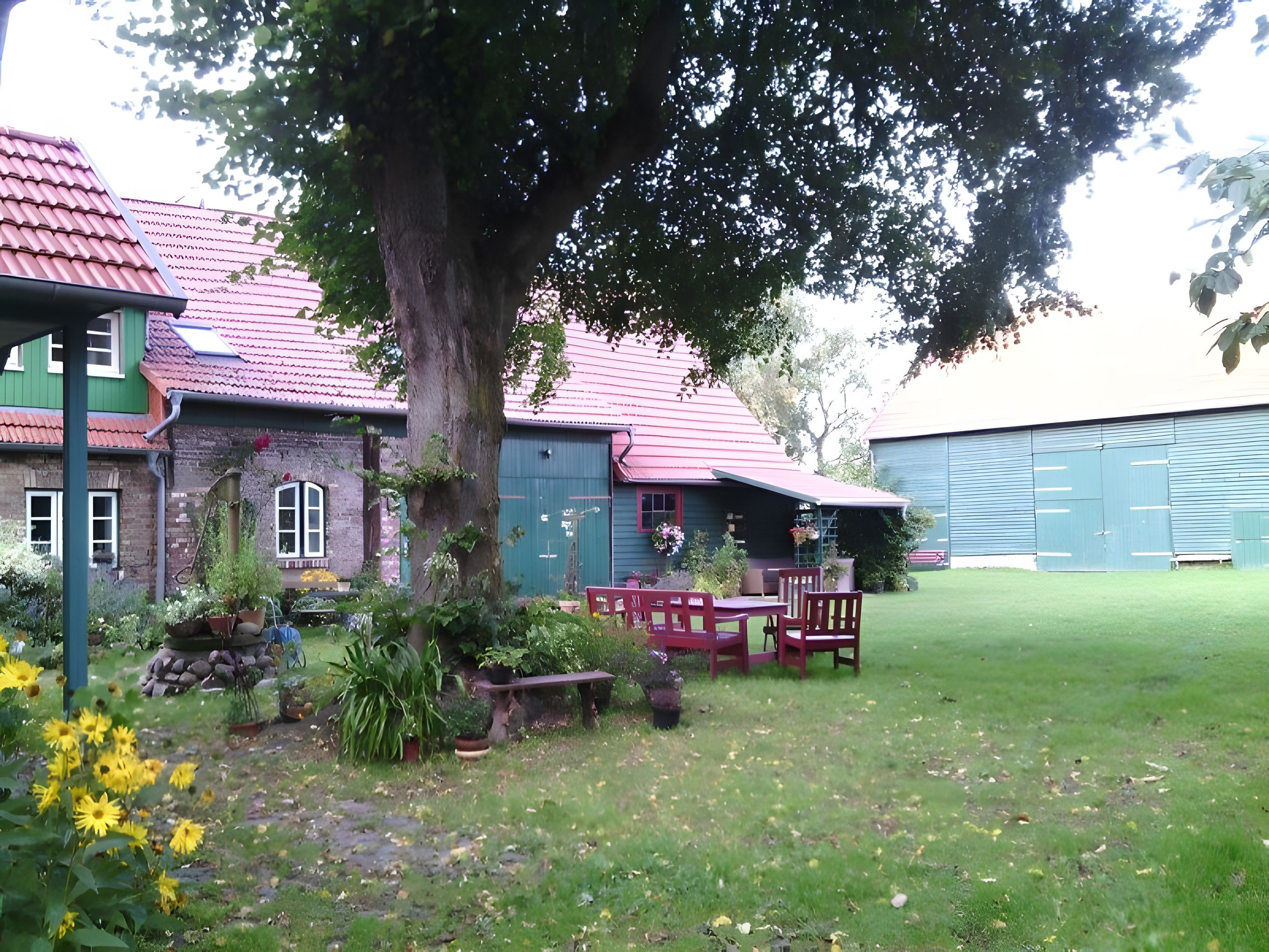 A building with a red roof, a large tree in front, and a grassy yard with a bench and chairs.