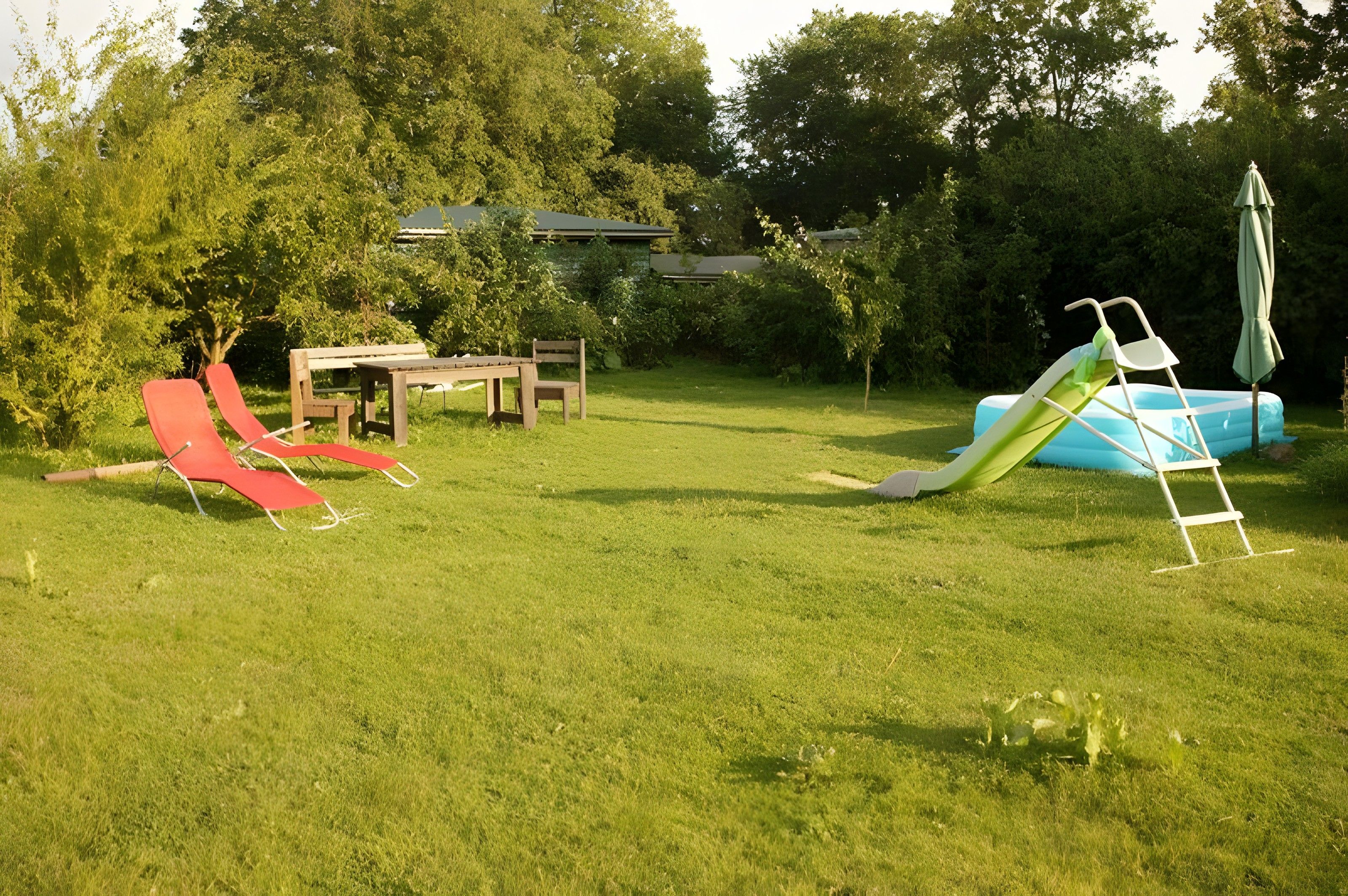 A grassy backyard with a red slide, a blue inflatable slide, a picnic table, and a green umbrella.