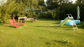 A grassy backyard with a red slide, a blue inflatable slide, a picnic table, and a green umbrella.