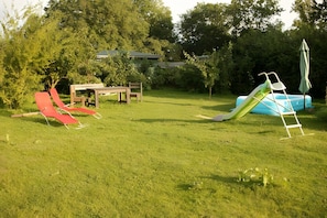 A grassy backyard with a red slide, a blue inflatable slide, a picnic table, and a green umbrella.