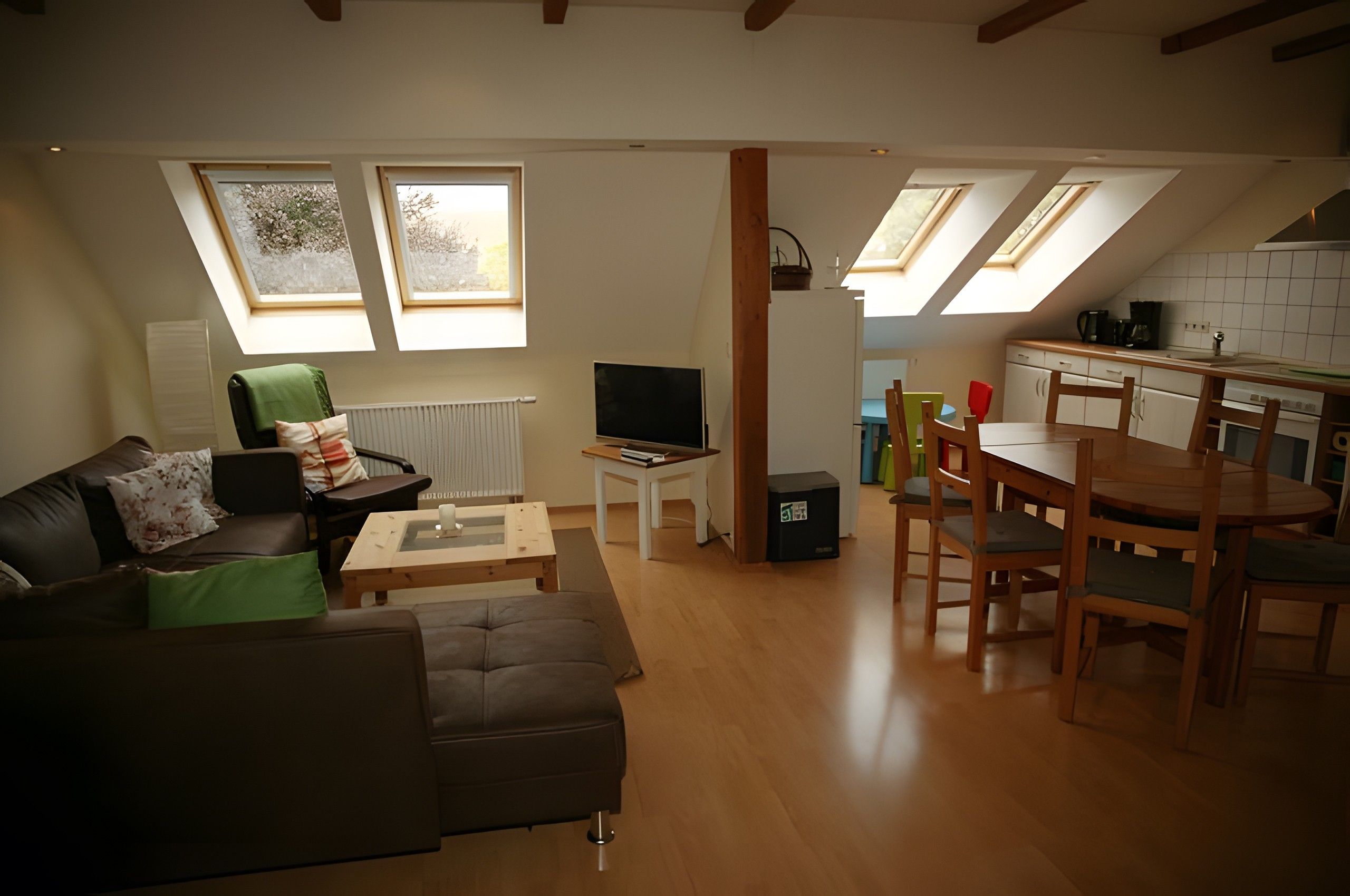 A modern living room with a sofa, a coffee table, a TV, and a dining area with a table and chairs. There are skylights above the kitchen area.
