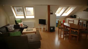 A modern living room with a sofa, a coffee table, a TV, and a dining area with a table and chairs. There are skylights above the kitchen area.