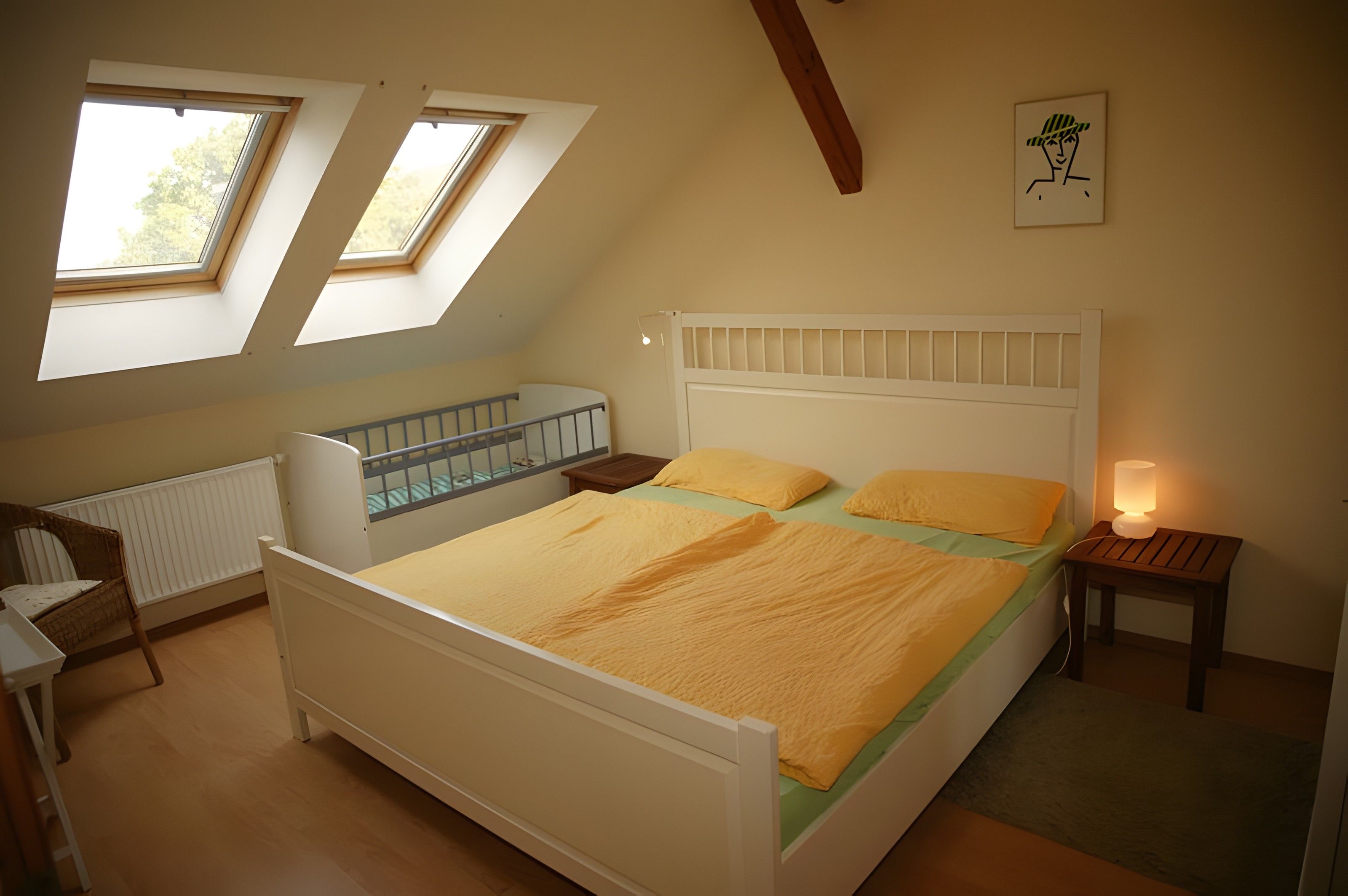 A bedroom with a bed, a crib, a nightstand, a lamp, and two windows.