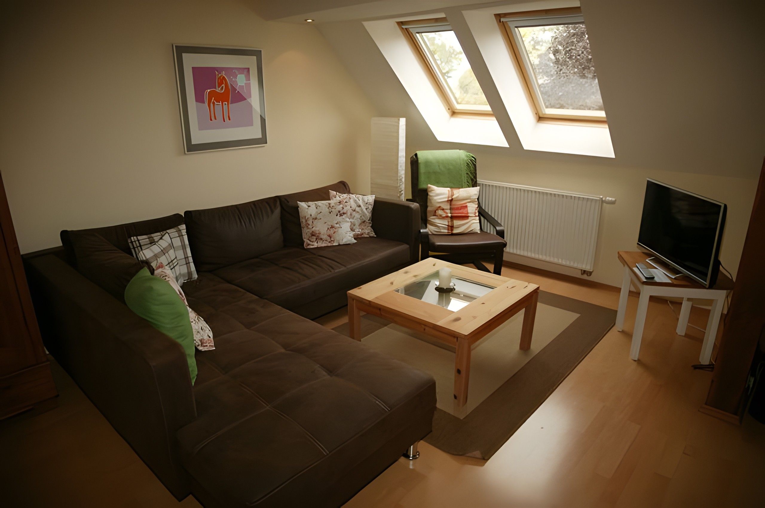 Ein Wohnzimmer mit einem braunen Sofa, einem Glastisch, einem grünen Sessel und einem Fernseher auf einem Beistelltisch.