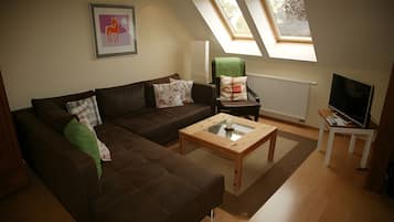 Ein Wohnzimmer mit einem braunen Sofa, einem Glastisch, einem grünen Sessel und einem Fernseher auf einem Beistelltisch.
