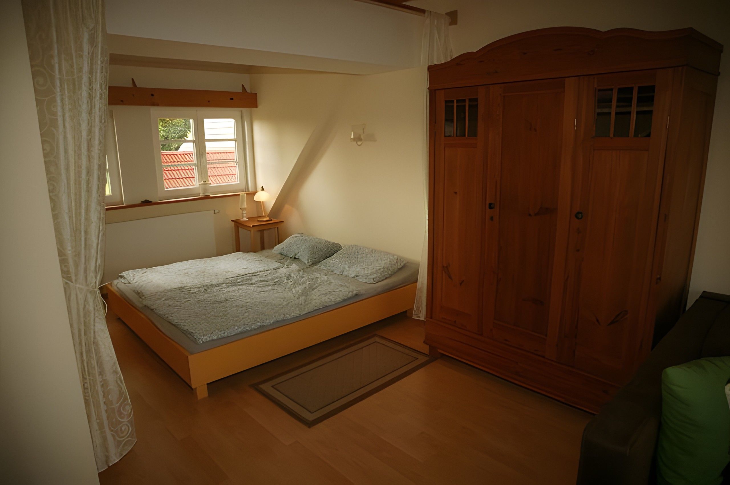 Un dormitorio con cama y armario de madera, una ventana con cortinas y una lámpara de noche.