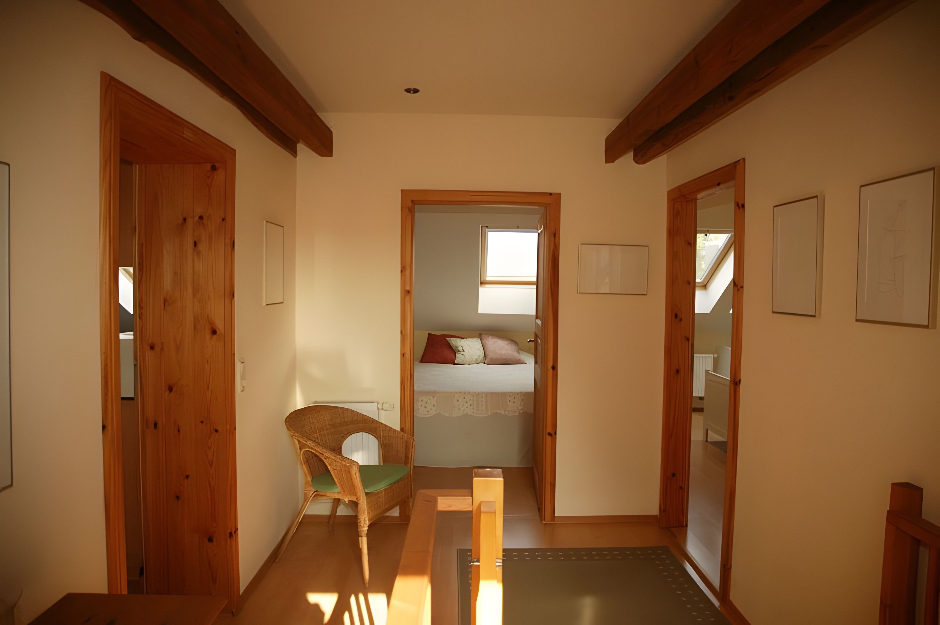 A narrow hallway with wooden doors, a wicker chair, a bed with a white bedspread and two pillows, and a skylight.