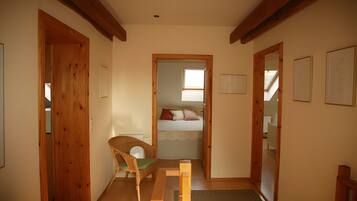 A narrow hallway with wooden doors, a wicker chair, a bed with a white bedspread and two pillows, and a skylight.