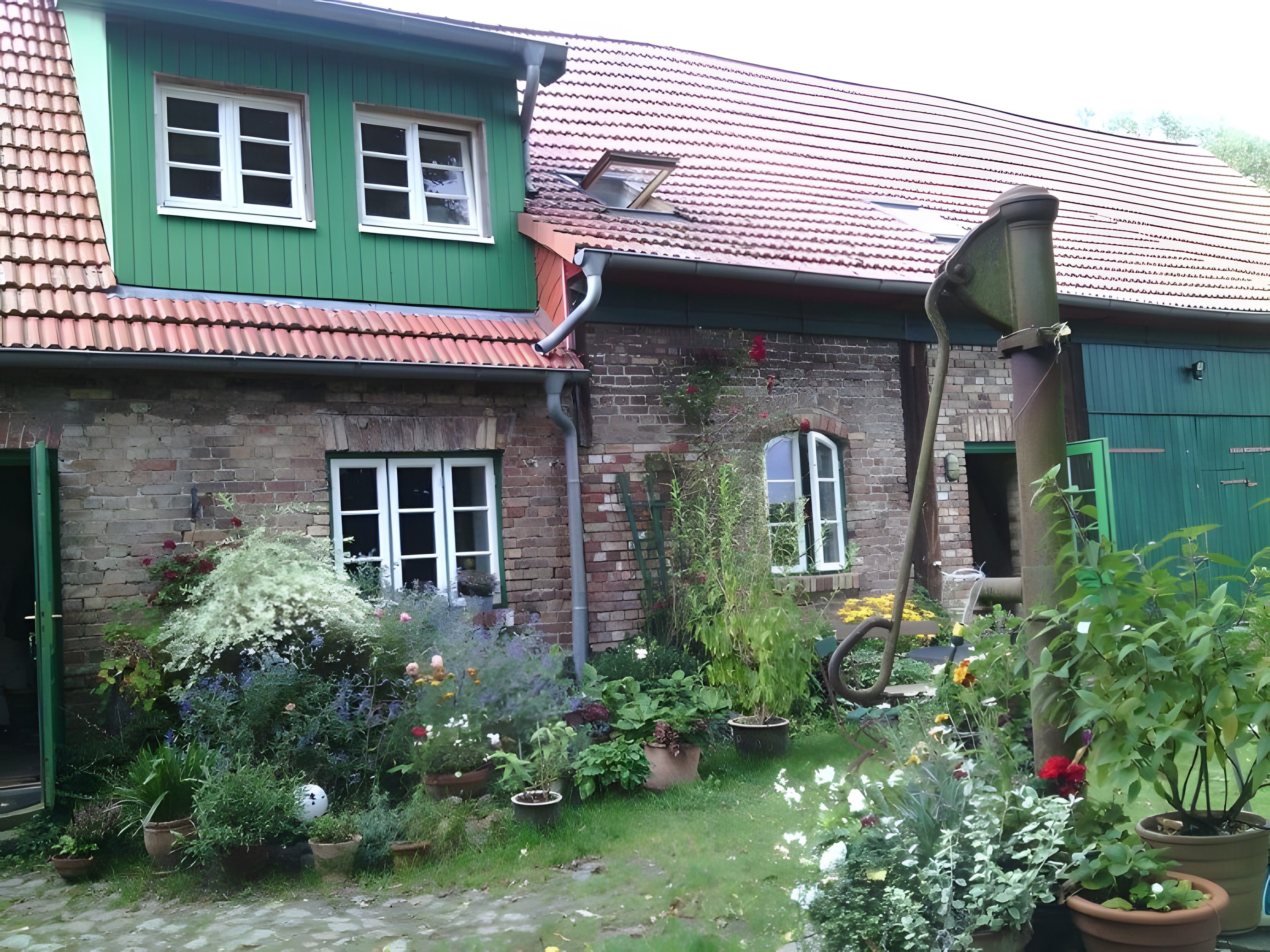 A brick house with green shutters, a tiled roof, and a garden with potted plants and flowers.