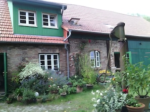 Ein Backsteinhaus mit grünen Fensterläden, einem Ziegeldach und einem Garten mit Blumentöpfen und Blumen.