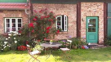 A brick house with a green door, a small garden with flowers, and a round table with chairs.