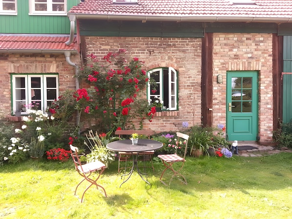Ein Ziegelhaus mit grüner Tür, ein kleiner Garten mit Blumen und ein runder Tisch mit Stühlen.