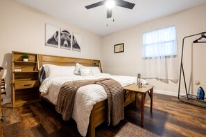 Un dormitorio con cama de madera, una mesita de noche, una lámpara en la mesita, una ventana con cortinas traslúcidas y un calentador de pared.