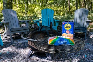 Ein Grill mit einer Tüte Marshmallows und einer Schachtel Honey Maid Cereal, umgeben von Adirondack-Stühlen in einem bewaldeten Gebiet.