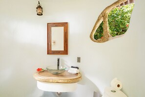 A bathroom with a glass bowl sink, a wooden-framed mirror, a wall-mounted light, and a small window with a plant. - Casa Verde – Oceanfront Beach House in Matapalo (Matapalo)