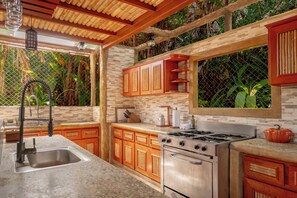 A kitchen with a stone backsplash, wooden cabinets, a stainless steel stove, and a window overlooking greenery. - Casa Aviari - Unique Rainforest Home in Matapalo (Matapalo)