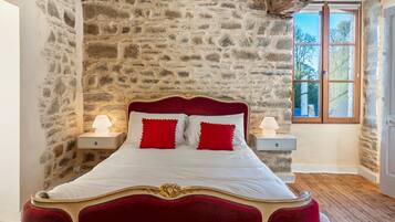 Un lit avec une tête de lit rouge et une literie blanche, flanqué de tables de chevet avec des lampes, dans une pièce aux murs en pierre et dotée d’une fenêtre.