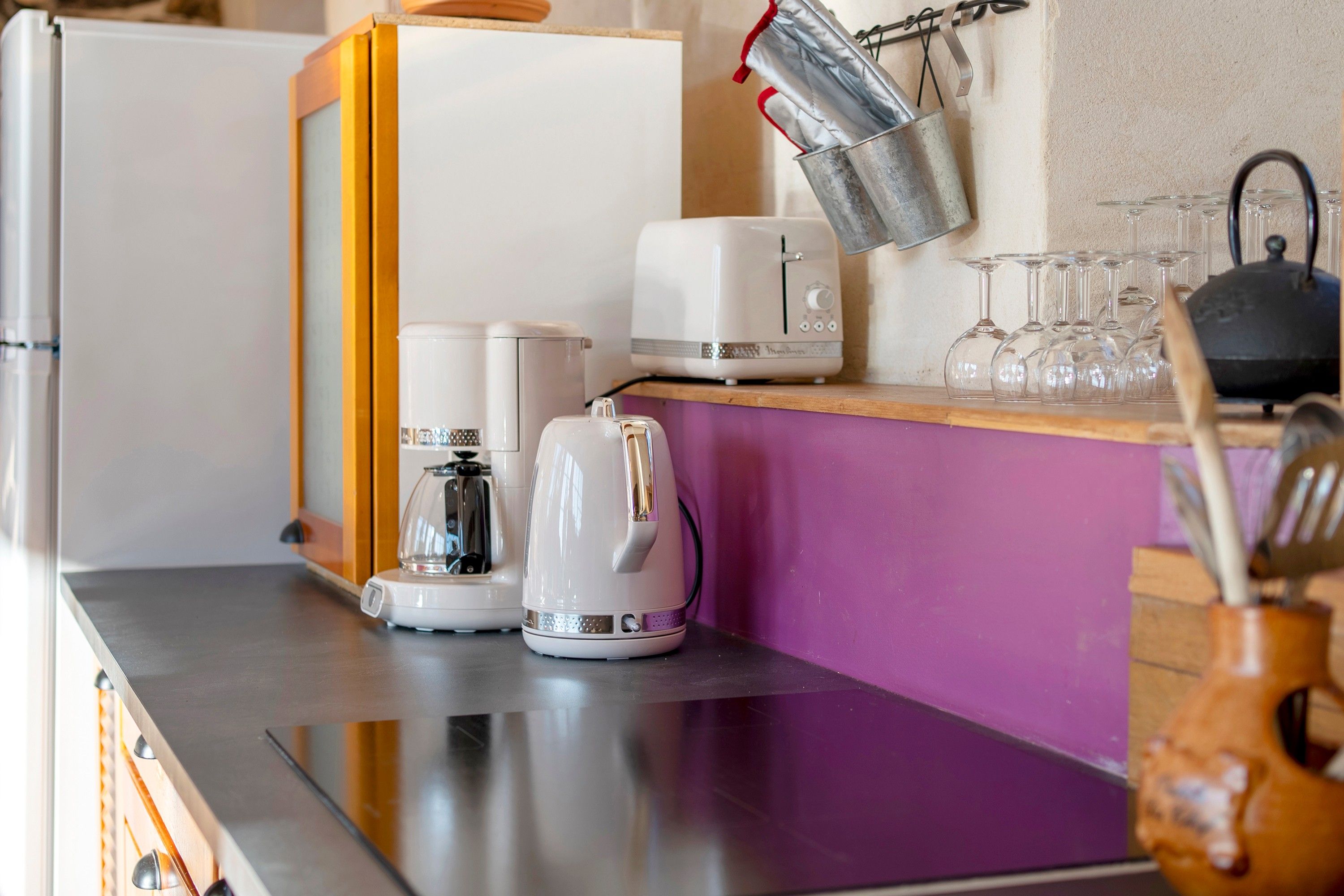 A kitchen with a toaster, kettle, and coffee maker on the counter, a hanging pot rack, and a shelf with wine glasses.
