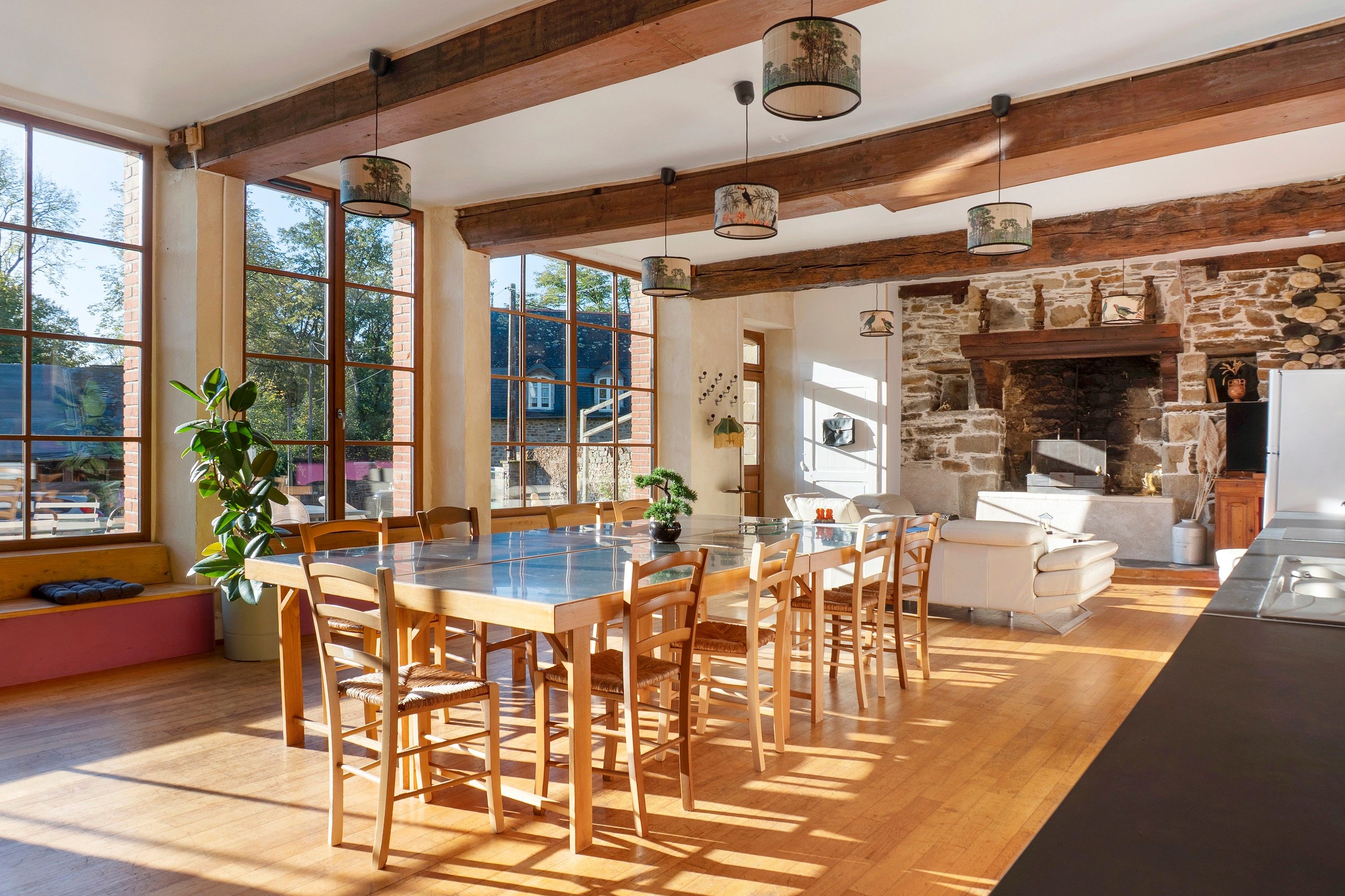 Une salle à manger spacieuse, dotée d’une grande table et de chaises, d’une cheminée en pierre et de grandes fenêtres laissant entrer la lumière naturelle.