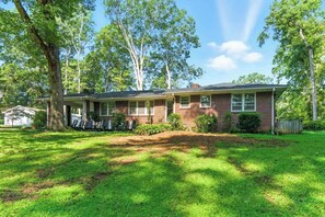 A single-story brick house with a covered porch, a lawn, and trees in the background. - Charming Ranch, 2BR Oasis Downtown Travelers Rest (Travelers Rest)
