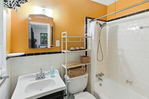 A bathroom with a white sink, toilet, and bathtub, a shelf with baskets, and a mirror with a decorative frame. - Charming Ranch, 2BR Oasis Downtown Travelers Rest (Travelers Rest)