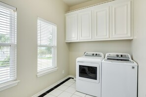 A laundry room with a front-loading washer and dryer, white cabinets above, and two windows with blinds. - Spacious Bayfront Retreat - Pet Friendly with Private Fishing Pier (Galveston)
