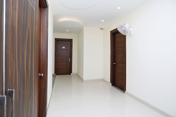 A hallway with a wooden door on the left, a white door in the center, and a white wall with a ceiling fan on the right. - Hotel O Royal Inn (Faridabad)