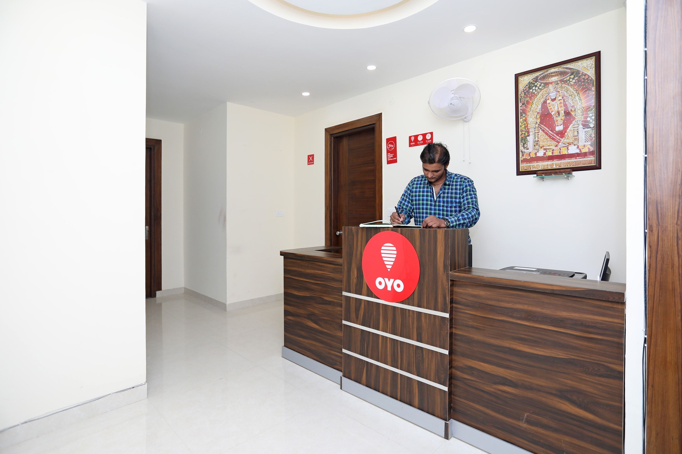 Bancada de recepção com logotipo, uma pessoa em pé atrás dela, um quadro na parede e uma porta ao fundo.