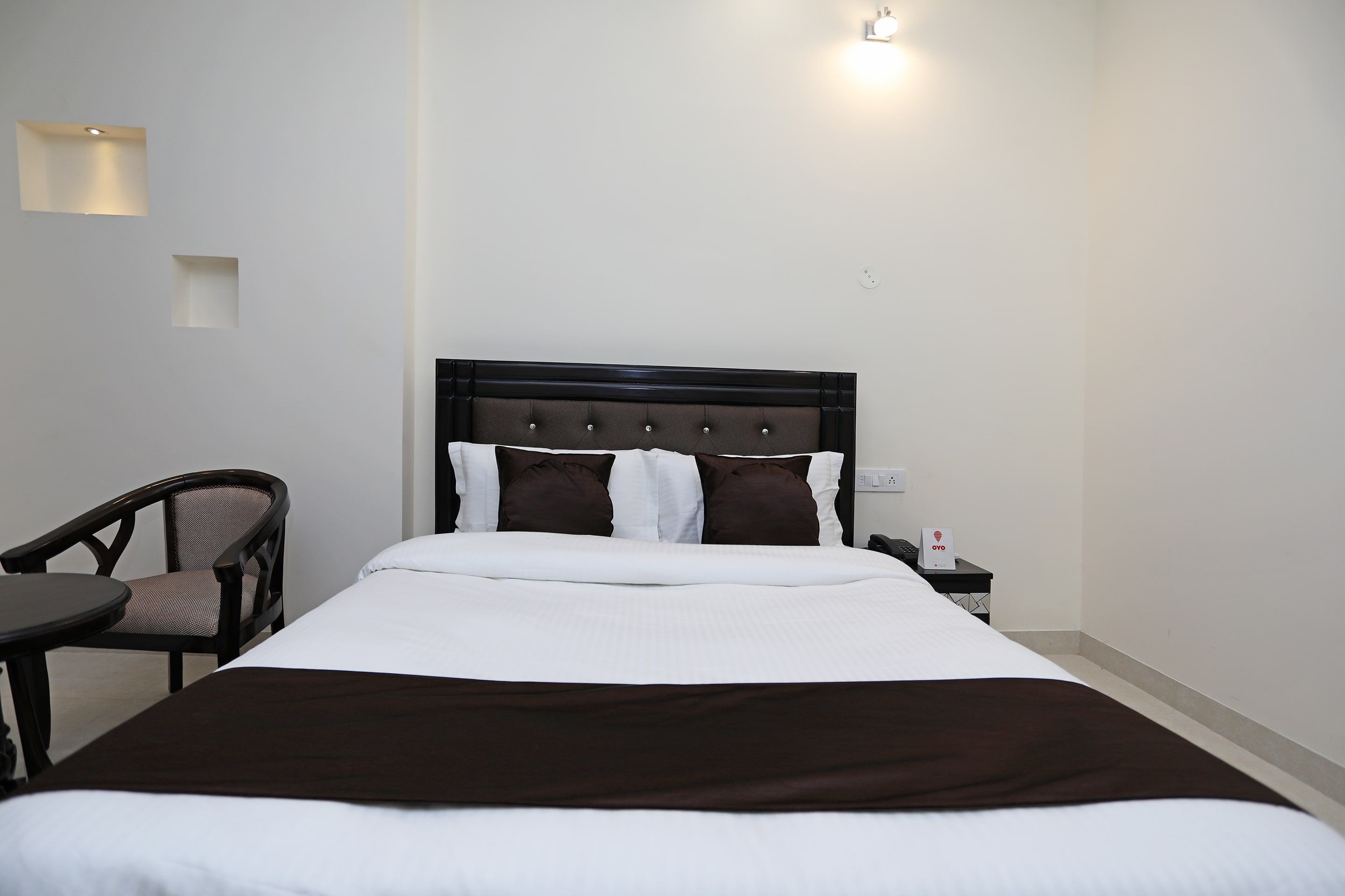 Quarto de hotel com cama, cadeira, mesa de cabeceira e luminária na parede.