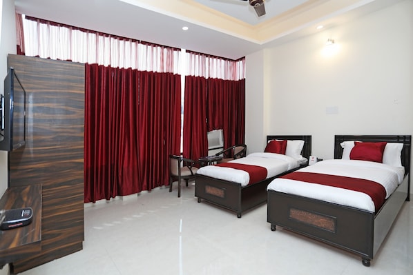 A hotel room with two beds, a desk, a chair, a TV, and a window with red curtains.