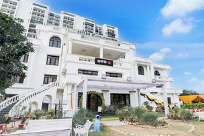 Um hotel com vários andares, entrada central, varandas e um jardim com plantas em vasos e áreas de estar.