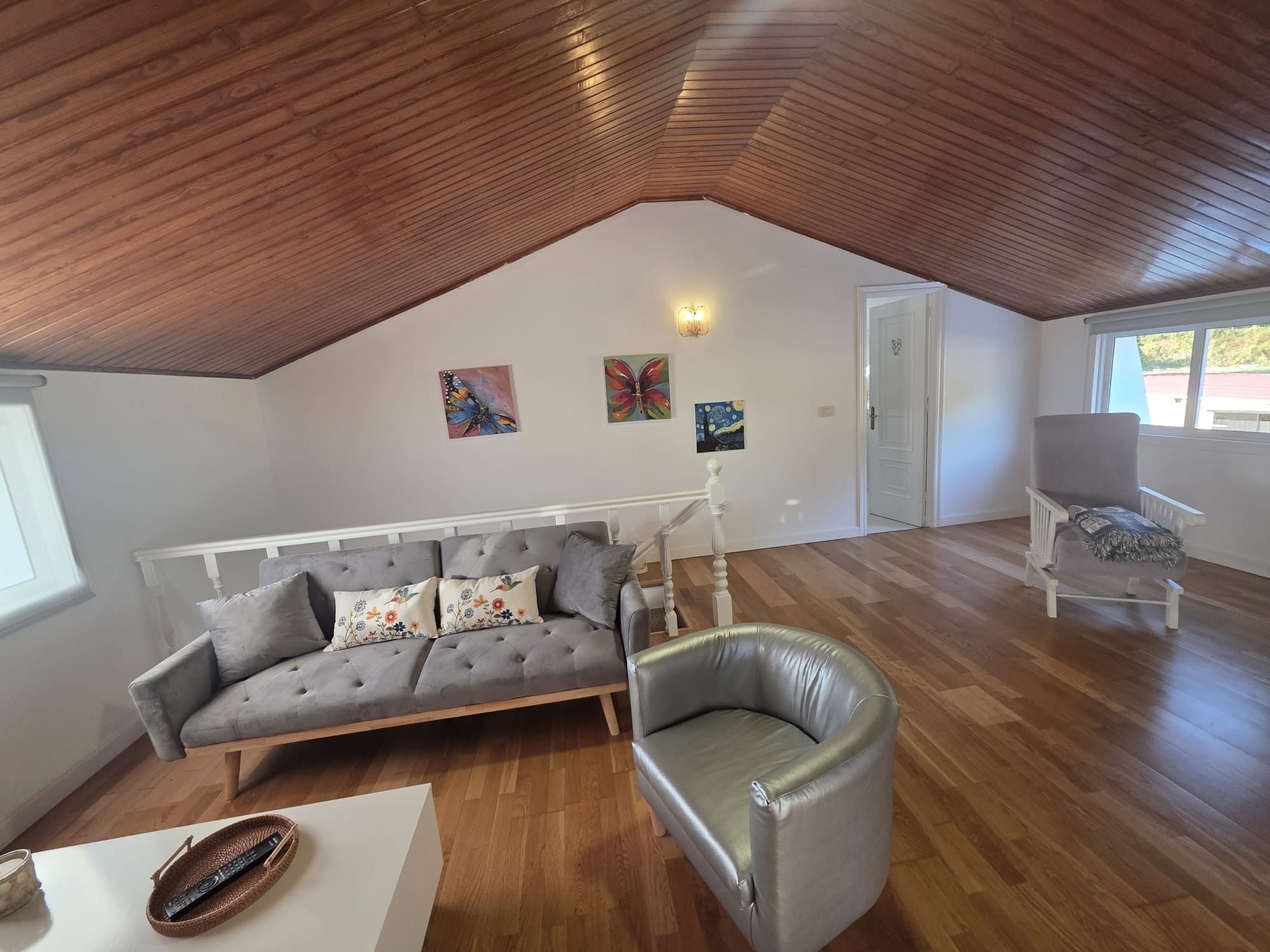 Un loft espacioso con un alto techo de madera, un sofá gris con cojines florales, un sillón de cuero y una puerta blanca que da al exterior.