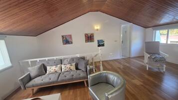 Un loft espacioso con un alto techo de madera, un sofá gris con cojines florales, un sillón de cuero y una puerta blanca que da al exterior.
