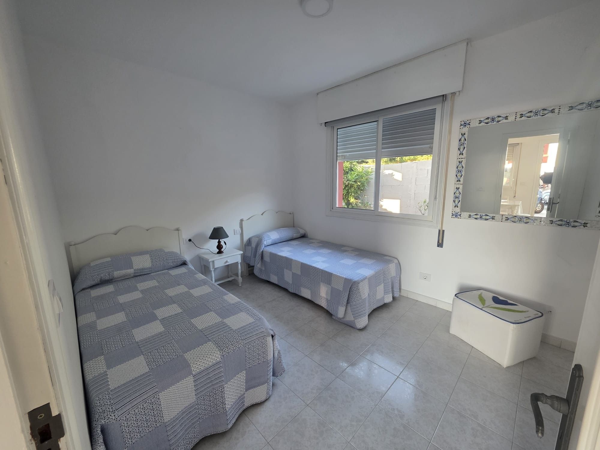 A bedroom with two beds, a bedside table with a lamp, a window with blinds, and a mirror on the wall.