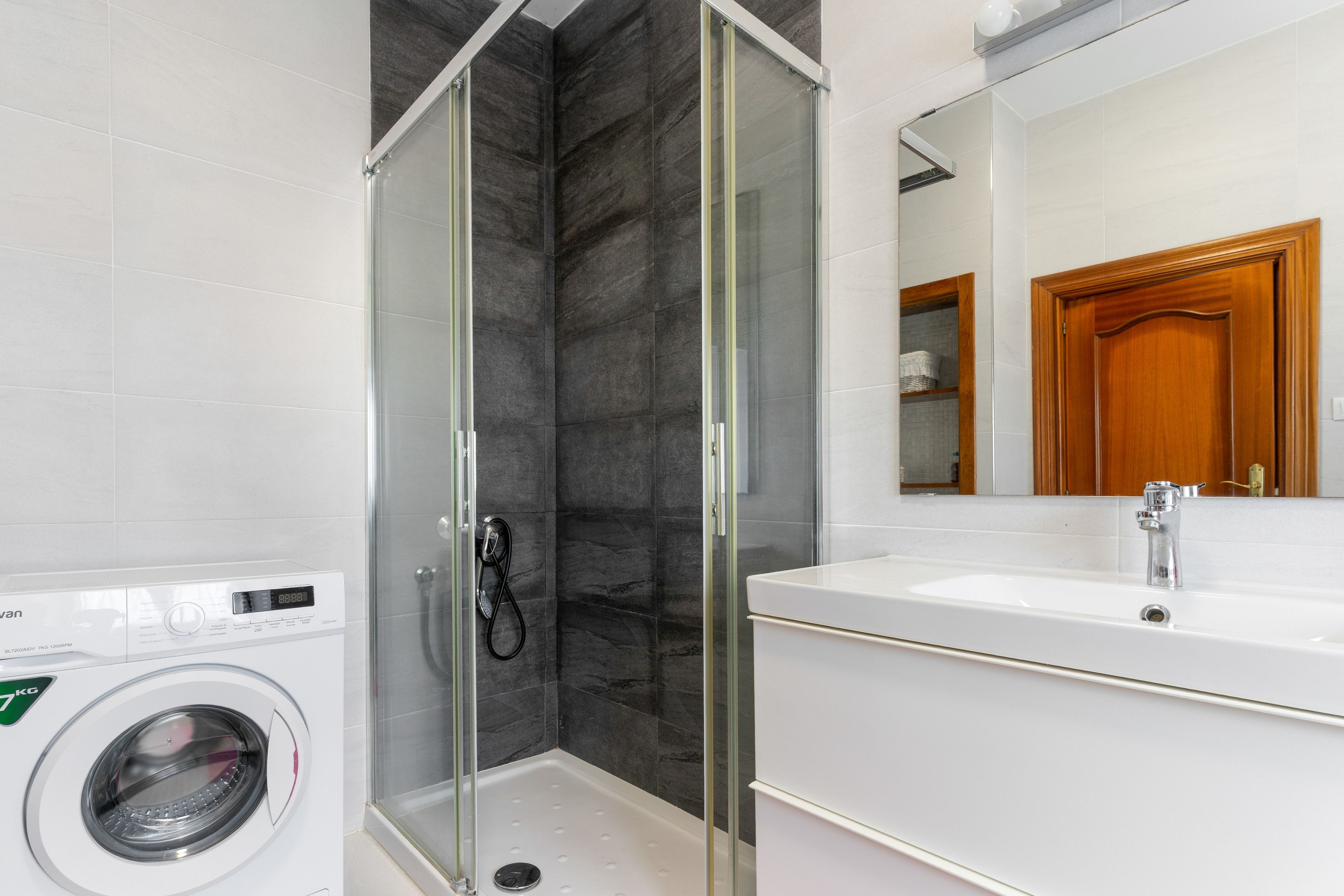 Un baño moderno con ducha de vidrio, lavamanos blanco y lavadora integrada.