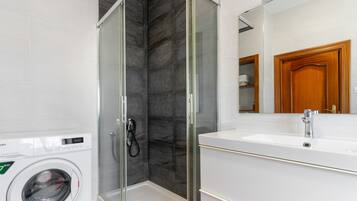 Un baño moderno con ducha de vidrio, lavamanos blanco y lavadora integrada.
