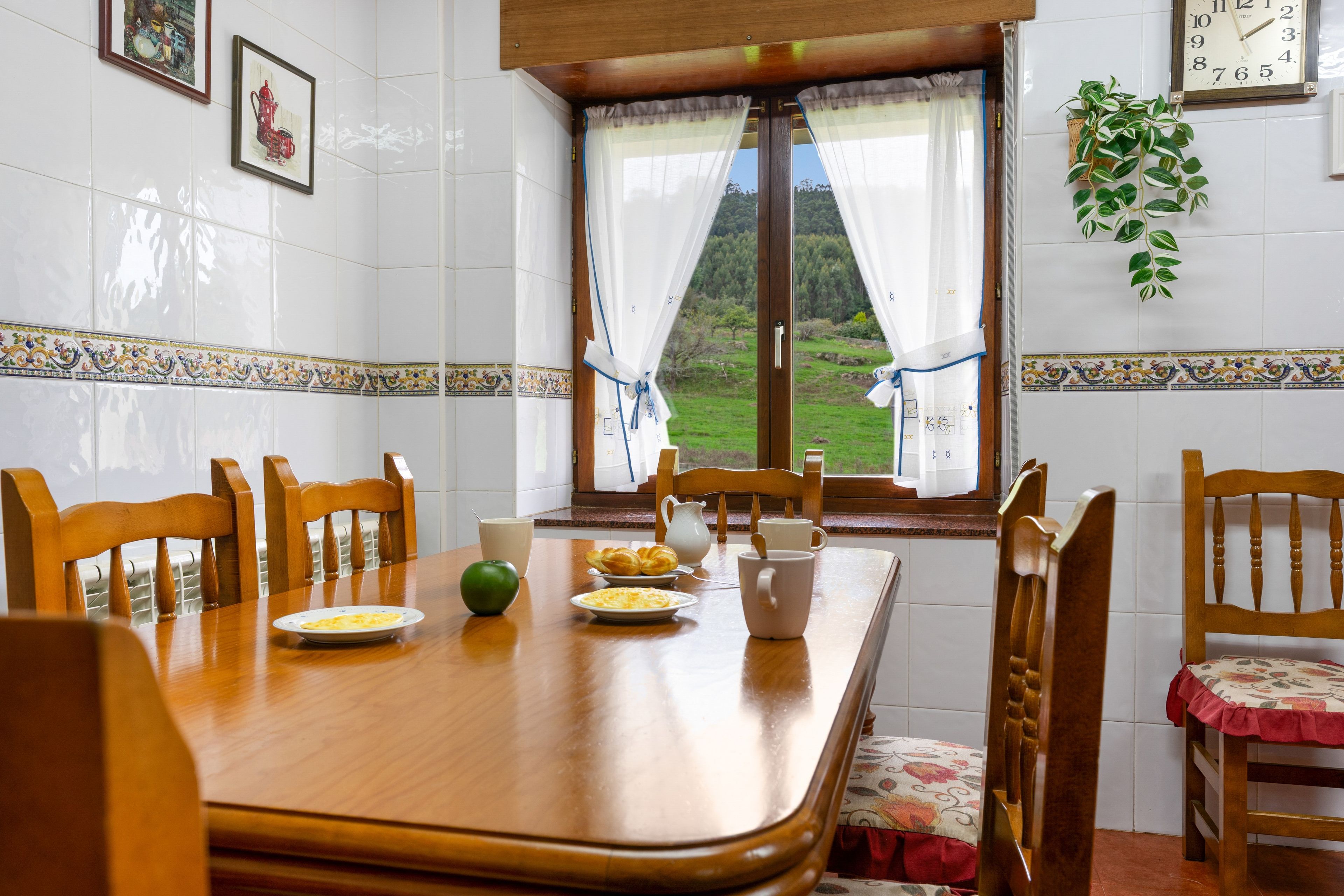 Un comedor con una mesa de madera, sillas y vistas a un paisaje verde a través de una ventana con cortinas.