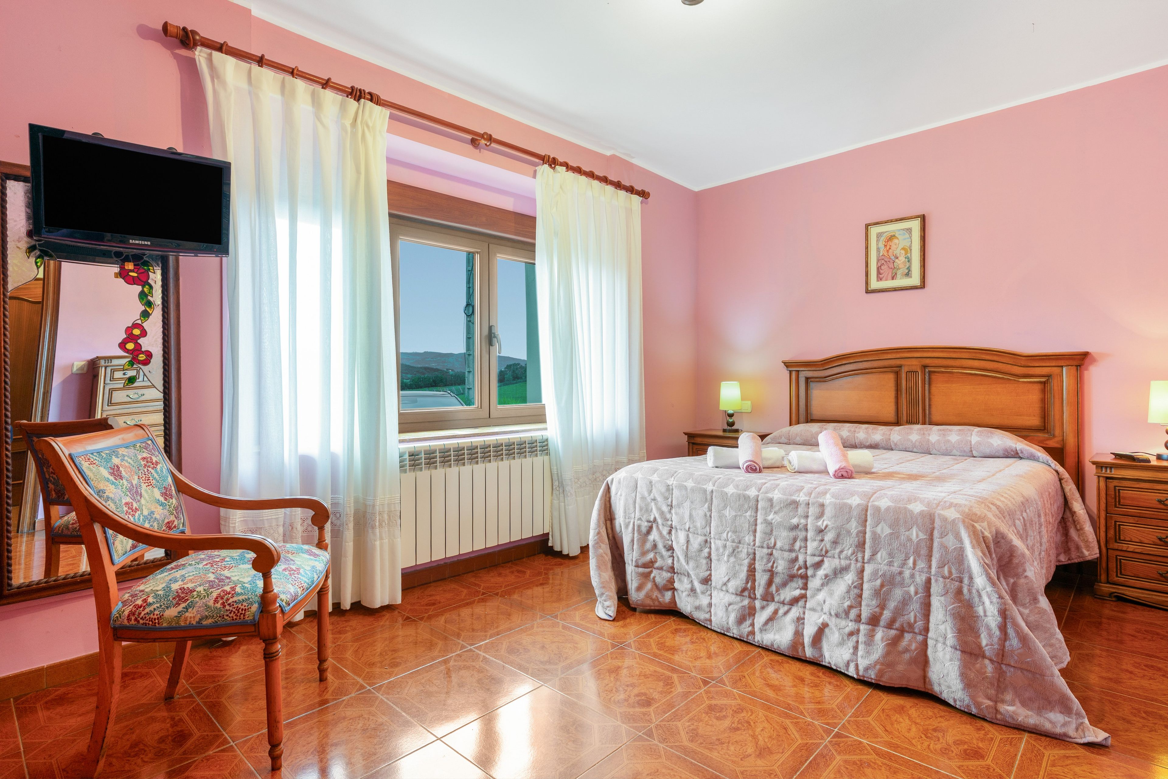 Un dormitorio con cama, una silla, ventana con cortinas, radiador y televisor montado en la pared.