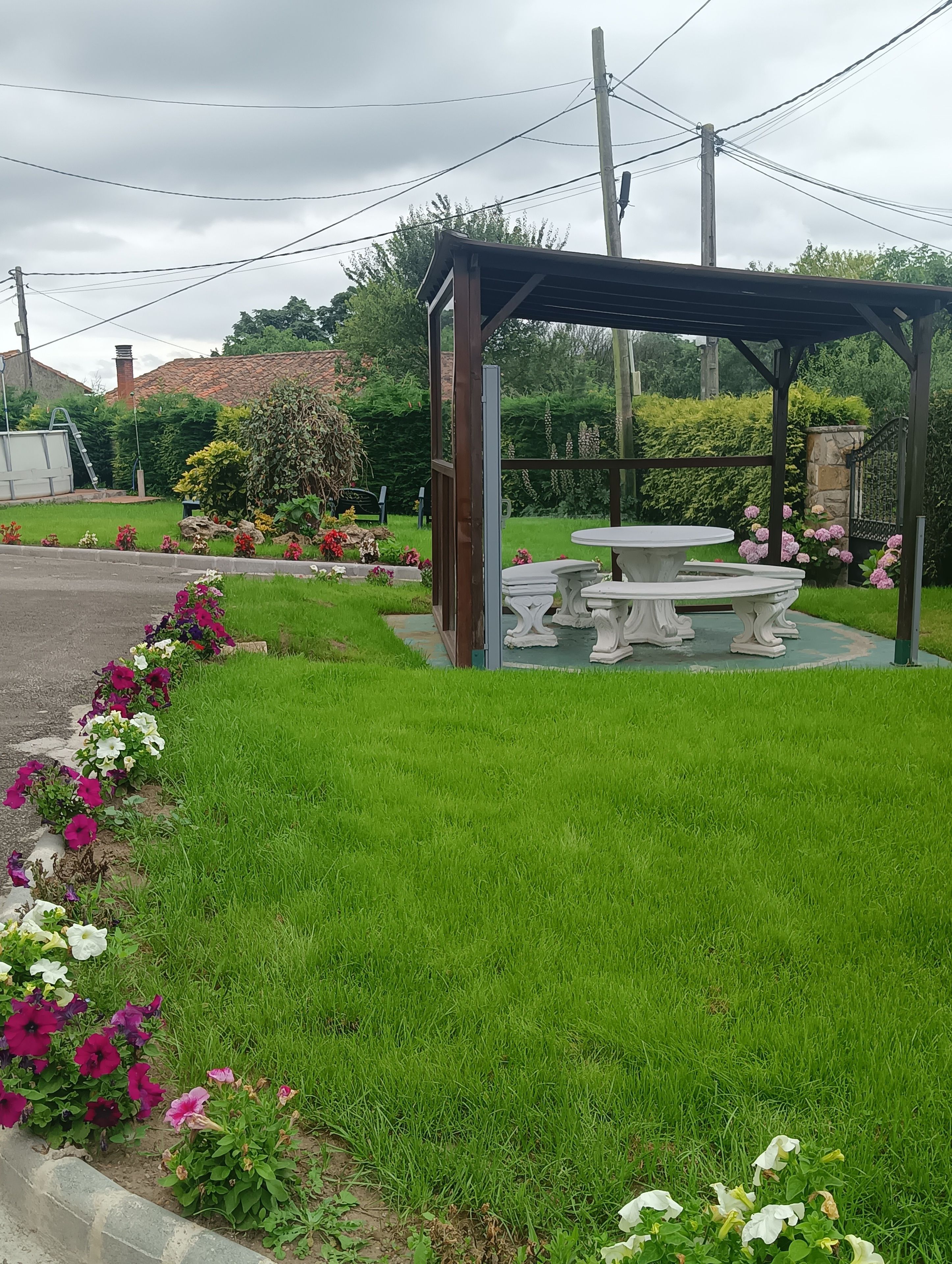 Un jardín con un pabellón, una mesa y bancos, rodeado de césped cuidado y macizos de flores.