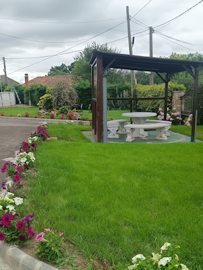 Un jardín con un pabellón, una mesa y bancos, rodeado de césped cuidado y macizos de flores.