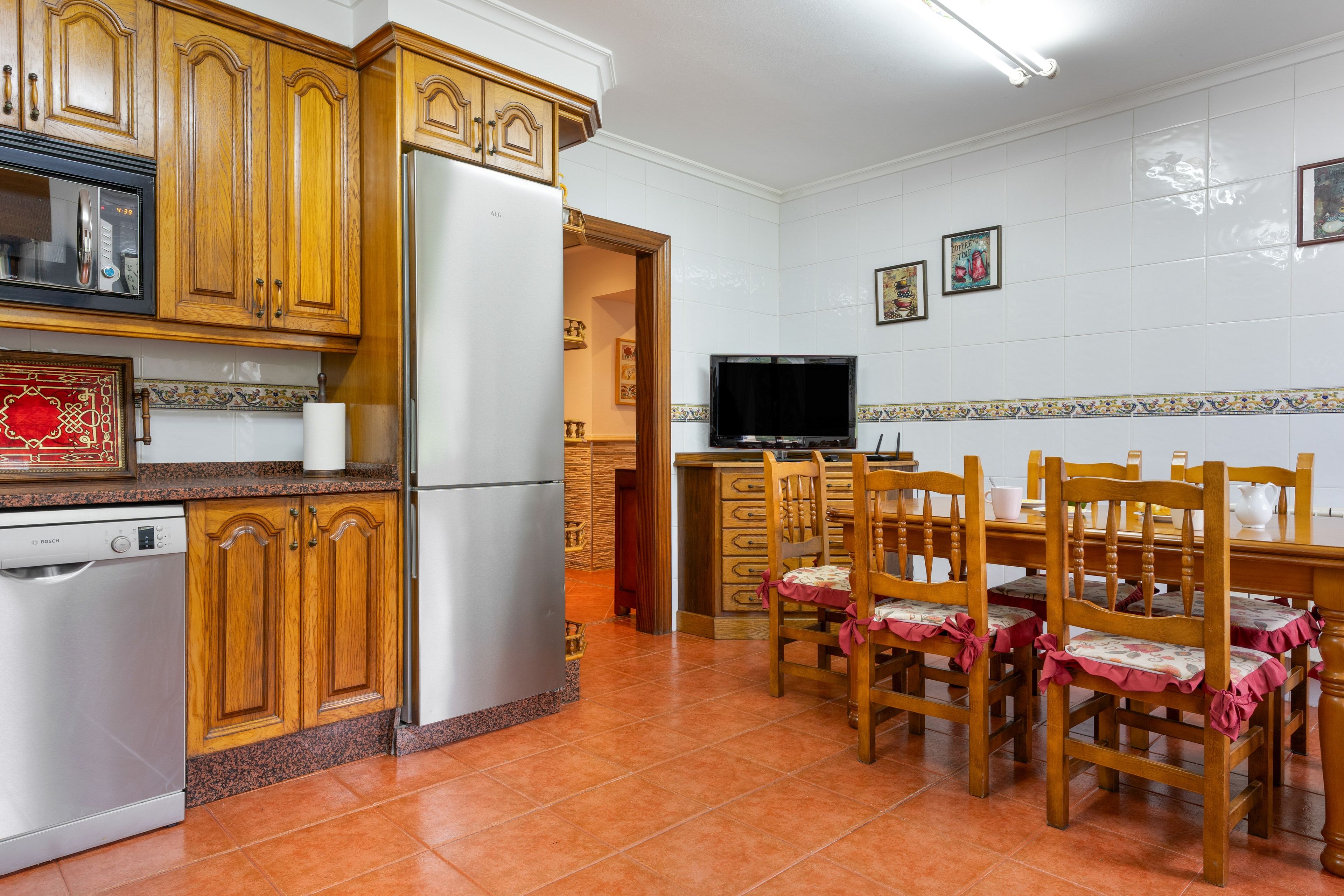 Une cuisine avec des meubles en bois, un réfrigérateur en acier inoxydable, un lave-vaisselle Bosch, une table à manger avec des chaises et un téléviseur.